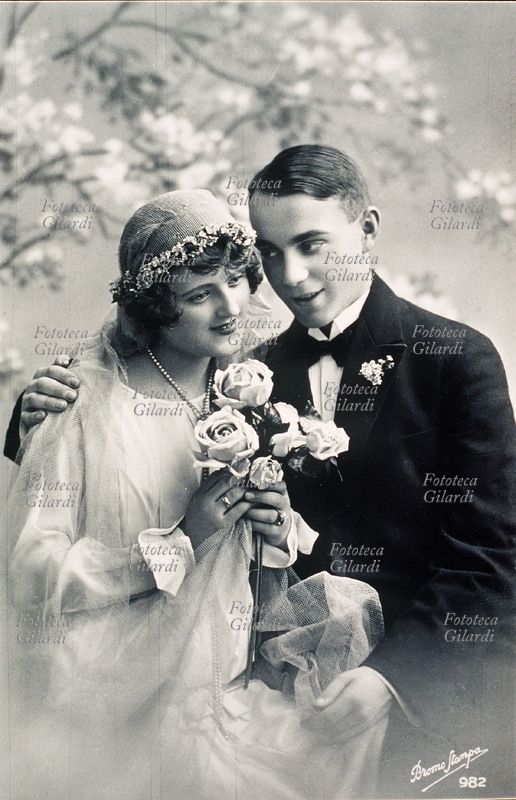 MATRIMONIO coppia di sposi ritratta a mezzobusto sullo sfondo di un albero dai fiori bianchi. La donna tiene un bouquet di rose chiare. Fotografia viraggio seppia; Bromo Stampa, Torino 1935