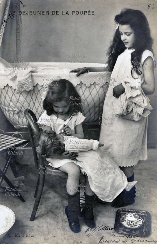 BAMBINI Il pranzo della bambola. Due bambine, in abito bianco, imboccano amorevolmente, una bambola con il cucchiaio. Fotografia viraggio seppia, Parigi 1902