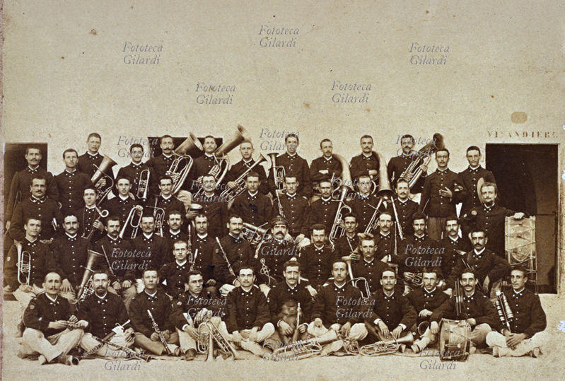 BANDA MILITARE Foto di gruppo ripresa sullo sfondo di un grande muro bianco, sulla porta a destra si legge "vivandiere". Al centro due graduati, a destra le percussioni, nelle prime file sulla sinistra i legni e poi lo spiegamento degli ottoni: in tutto 51 elementi. I due commilitoni seduti a terra sulla sinistra condividono con un gesto simbolico un grappolino d\