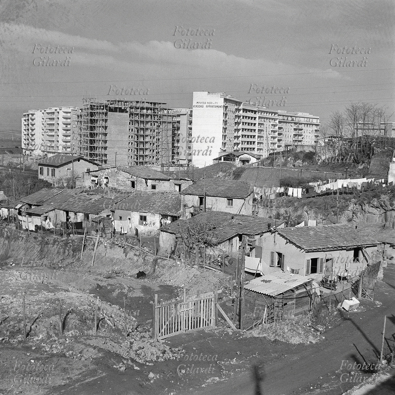 EDILIZIA boom economico: nuove case in costruzione alla periferia della città, in estremo contrasto accanto al quartiere povero di casette fatiscenti. Sulla facciata del condominio più alto si legge: \