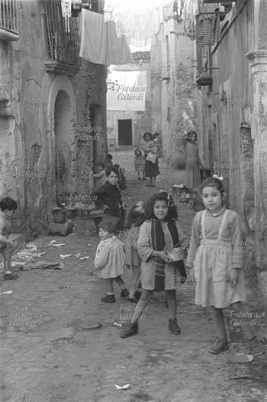 BAMBINI di varie età giocano in strada. Una bimba ha in mano un pentolino. Sullo sfondo una mamma con in braccio il bambino più piccolo assiste alla scena. Situazione di degrado e sporcizia, tuttavia i bambini sono paffuti e puliti. Fotografia di Ando Gilardi (1921 - 2012) #andogilardi, Crotone 1957.