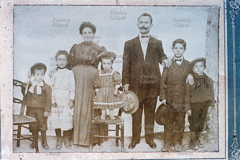 FAMIGLIA genitori in posa con i cinque figli, tre maschi e due femmine. Stanno tutti in piedi, e la più giovane e più piccola di statura posa in piedi sopra una sedia, mentre il maggiore porta già i calzoni lunghi e il cravattino, e tiene il cappello in mano come il papà. Fotografia di anonimo eseguita in un atelier ambulante, Italia 1890 circa.