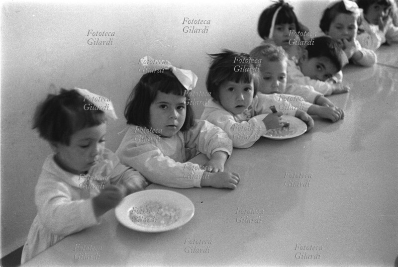 BAMBINI di scuola materna attendono, seduti ai banchi del refettorio, il pranzo. Ad alcuni di loro le suore hanno già servito un bel piatto di minestra. Tutte le bambine recano in testa un fiocco bianco. Vari fotogrammi (Lucania, San Costantino Albanese, Spedizione De Martino 1957). Fotografia di Ando Gilardi (1921 -2012) #andogilardi, Italia 1957.
