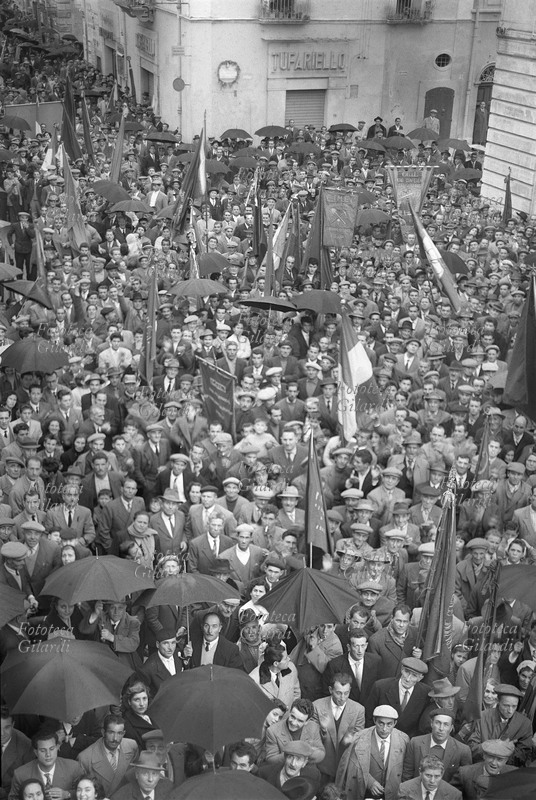 Giuseppe DI VITTORIO (1892-1957). Folla commossa a Cerignola alla manifestazione per la prima commemorazione della morte. Qualcuno porta la bandiera rossa, altri il tricolore o uno stendardo, molti si riparano con l\
