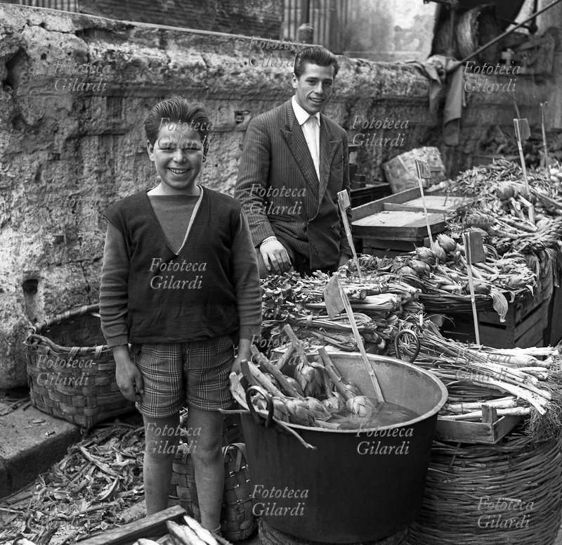 Smiling CHILD at work together with a young family member, at the vegetable stand where artichokes, celery and spring onions are displayed. Photograph by Ando Gilardi (1921 - 2012) #andogilardi, Palermo circa 1955.