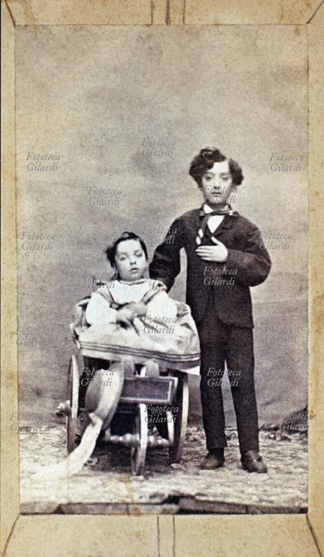 BAMBINI Due fratelli (due e sette anni circa) posano per una fotografia ricordo. Il grande è in piedi con elegante vestito scuro. Il piccolo è seduto nel passeggino ottocentesco, con tre ruote: due grandi laterali e una piccola centrale anteriore. Italia, 1880 circa.