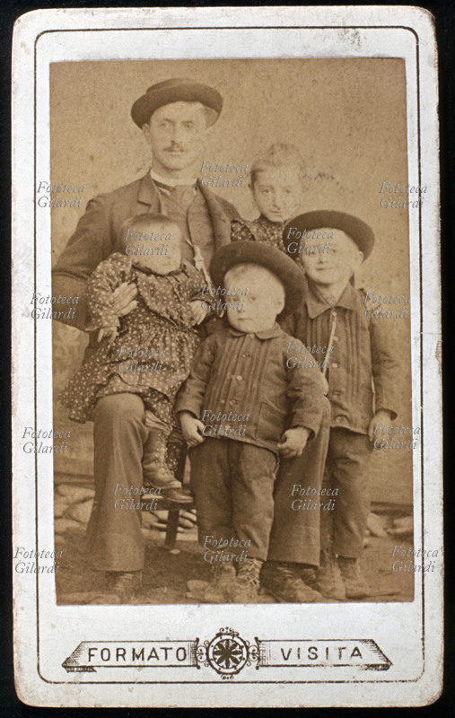 FAMIGLIA giovane padre e quattro figli, due maschi e due femmine, tutti fermi e con espressione serena tranne la più giovane, in braccio al papà, visibilmente irrequieta. Le due femmine sono vestite uguali, così come i maschi che portano il cappello come quello di papà. Fotografia in formato \