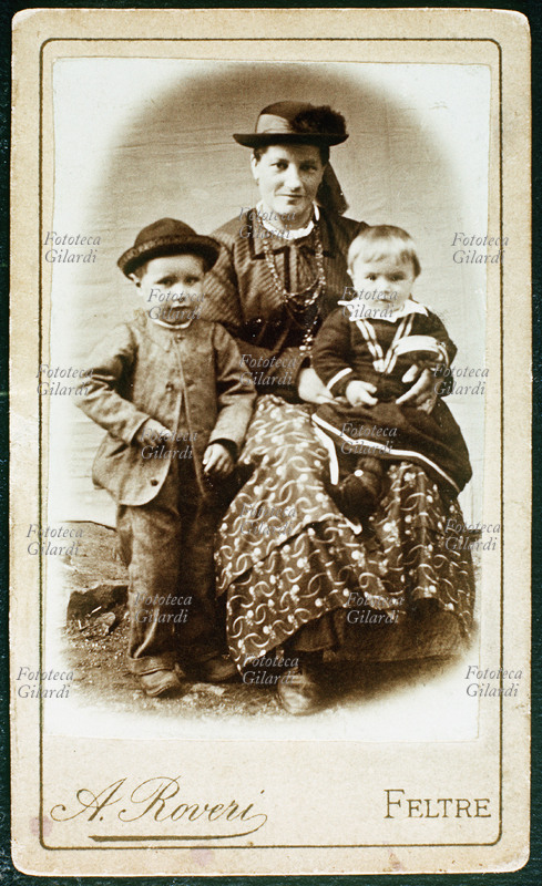 FAMIGLIA Ritratto fotoricordo di madre con due figli (uno e tre anni). La madre tiene in grembo il più piccolo e indossa il costume tradizionale trentino. Studio fotografico A. Roveri, Feltre 1890 circa.