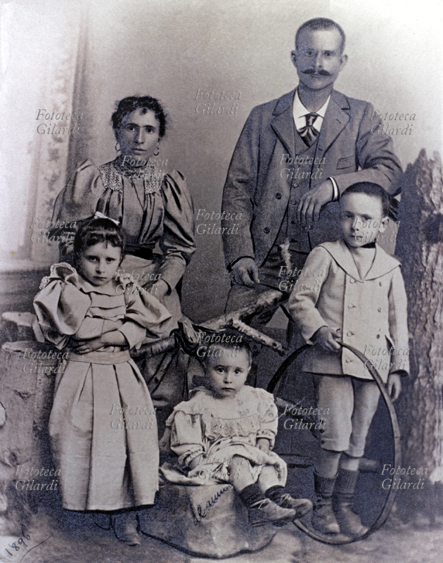 FAMIGLIA Genitori in posa con tre figli. I tre bimbi sono in primo piano, da sin: la secondogenita in abito con maniche a sbuffo molto elegante; il più giovane (2 anni) seduto su un masso; il primogenito con il gioco del cerchio. Italia 1910 circa