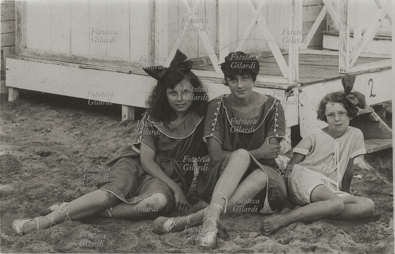 VACANZE, tre adolescenti, probabilmente sorelle data la somiglianza, posano per una foto ricordo al mare sedute sulla sabbia vicino alla piattaforma delle cabine. Italia, 1910 - 1920