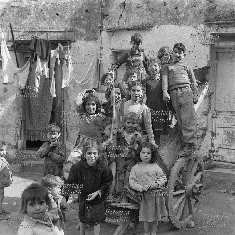 BAMBINI vivaci, in posa sopra un carretto in un vicolo del quartiere. Molti hanno espressioni scherzose e spavalde. Solo un piccolo, tenuto per mano dalla sorella più grande, piange. Fotografia di Ando Gilardi (1921 - 2012) #andogilardi da ampio servizio, Palermo, Cortile Cascino, 1957.