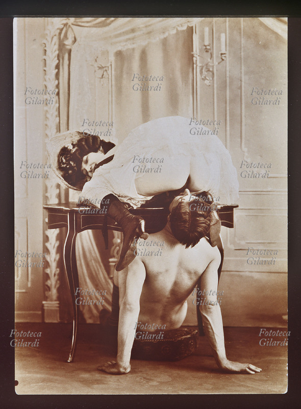 CUNNILINCTUS acrobatico eseguito da una spiritosa coppia di amanti: lei vestita di pizzo bianco e cappellino floreale, alza la gonna accucciata su un piccolo scrittoio e offre al compagno l\