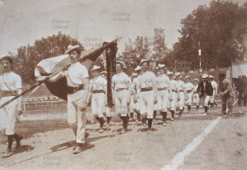 SPORT group of athletes running into an athletics stadium, parading under the Italian national flag. Photograph, Italy, circa 1900.