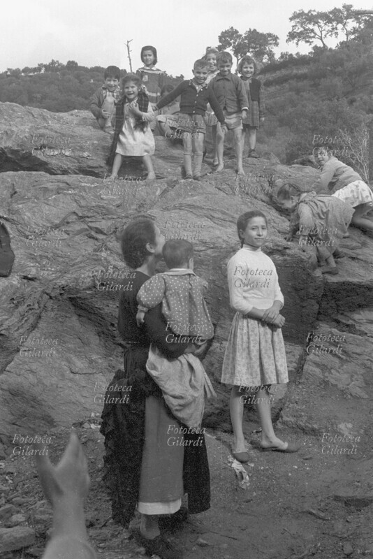 BAMBINI di diverse età giocano in una campagna rocciosa divertendosi molto. La madre li accompagna con il fratellino più piccolo in braccio. Fotografia di Ando Gilardi (1921 - 2012) #andogilardi, Nicastro 1955-58.