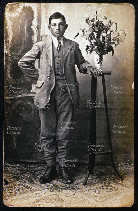 RITRATTO maschile a figura intera. Il ragazzo, ancora imberbe, indossa un elegante completo grigio con panciotto uguale, camicia bianca e cravatta, accessori con nodo a triangolo con grandi disegni. Fotografia d\