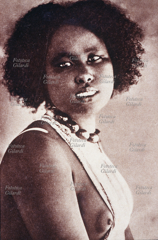 COLONIALISMO A.O.I./Somalia. Donna somala, in posa di tre quarti, con un abito che le scopre il seno. Cartolina litografica, Mogadiscio s.d.
