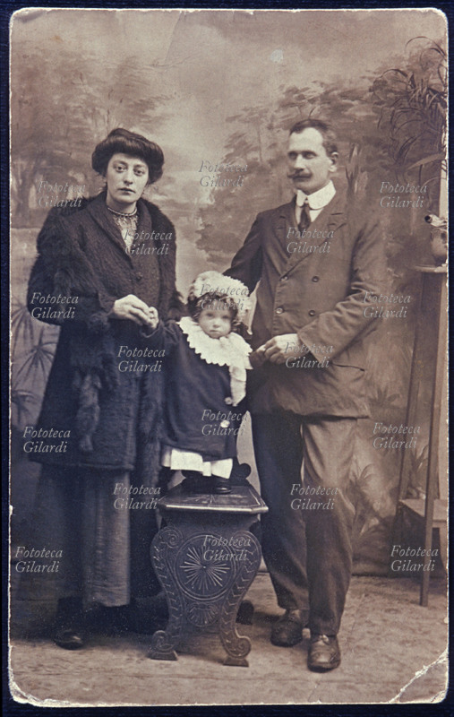 FAMIGLIA genitori eleganti e figlia bambina, ricciuta, bionda, fotografata in piedi sopra uno sgabello tra il padre e la madre; la tengono ferma per la lunga posa, tuttavia la piccola non rimane perfettamente immobile. La madre indossa un abito lungo scuro, un soprabito di maglia con una lunga stola di pelliccia. Fotografia di anonimo, eseguita in atelier. Italia, 1910 circa.