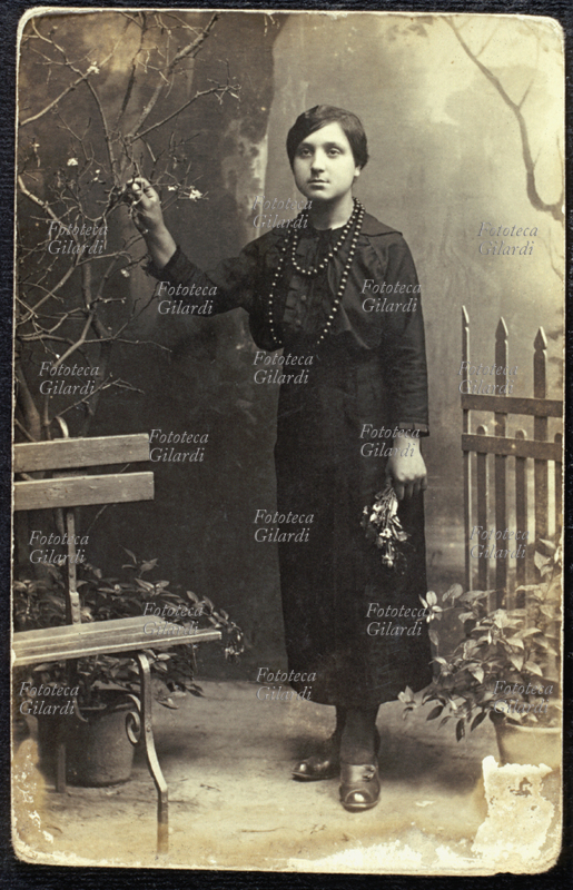 MODA Ritratto di giovane donna a figura intera con vestito scuro. La posa è realizzata in uno studio fotografico, ma bene ambientata: è stata ricreata con un fondale e pochi elementi (una panchina, rami secchi, vasi di fiori e un cancelletto) una veduta che simula un esterno. Italia, 1900 circa.