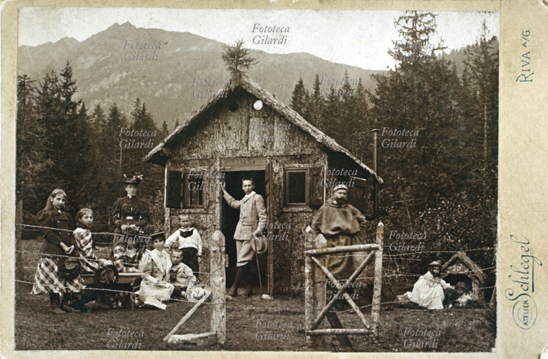 VACANZE MONTAGNA Bambini e giovanetti, amici e parenti, accompagnati da una governante ed un frate, sostano in una curiosa casetta di legno nella radura di una pineta montana. Le due bimbe più piccole, per gioco, dividono la cuccia con il cane. Fotografia virata seppia, s.d.