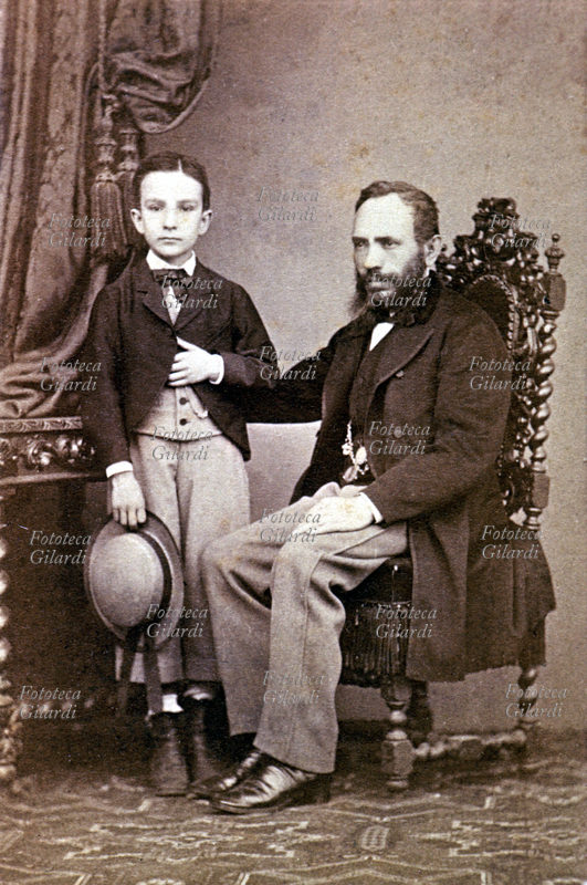 FAMIGLIA Padre e figlio di circa 10 anni posano per una fotografia ricordo. Sono vestiti in modo elegante, anche il bambino ha una giacca scura e i calzoni chiari intonati al gilet; in una mano tiene una paglietta con fiocco, l\