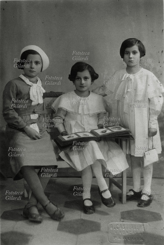 BAMBINI Tre sorelle in posa per una fotografia ricordo: due sono sedute e una è in piedi; quella al centro tiene appoggiato sulle ginocchia l\