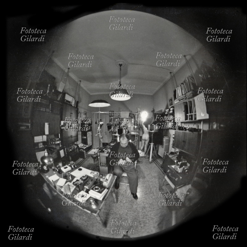 Ando GILARDI (1921-2012) autoscatto con fish-eye che lo ritrae nel suo laboratorio di Roma mentre sta mettendo a punto il suo apparecchio per effettuare una seduta fotografica di nudo; sullo sfondo la modella si prepara alle pose. Roma, 1964 circa.
