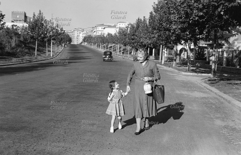 FAMIGLIA nonna e nipotina tornano a casa dalla spesa, chiacchierando e tenendosi per mano in un viale alberato di Roma. La signora porta sul braccio una borsa che visibilmente contiene i generi alimentari appena acquistati e con la mano regge un fiasco di vino. Le due camminano ignorando il marciapiede, in una strada dal traffico quasi inesistente; si vede solo una automobile allontanarsi in direzione opposta. Fotografia di Ando Gilardi (1921-2012) #andogilardi, Roma 1955 circa