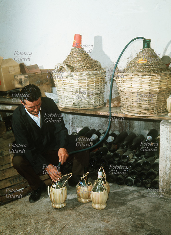 VINO Imbottigliamento di vino in una cantina privata. Con il tradizionale metodo dei vasi comunicanti il vino viene trasferito dalla damigiana ai fiaschi. Fotografia di Ando Gilardi (1921-2012) #andogilardi, Italia 1960 circa.