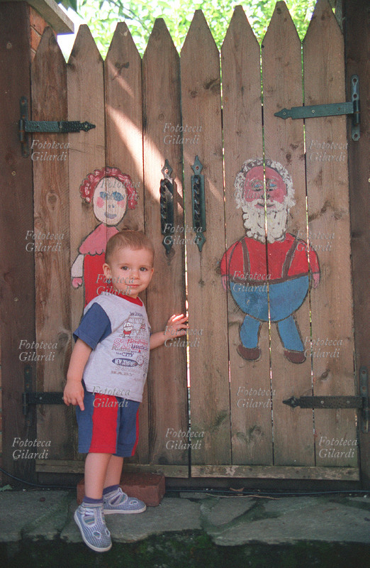 BAMBINI Un bimbo sale i gradini verso un cancelletto con le figure degli antenati che raffigurano insieme, il suo passato e il suo divenire. Il suo percorso va verso il futuro, ma guarda timidamente indietro per essere rassicurato. Caldasio (AL), Agosto 2000. Fotografia di Elena Piccini