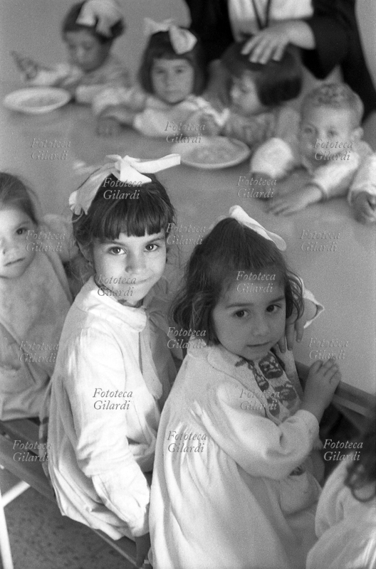 BAMBINI Bambine della scuola materna attendono il pranzo, sedute ai banchi del refettorio. Indossano il bavaglino per proteggere i grembiulini bianchi, e quasi tutte un nastro bianco fra i capelli. Immagine presa durante la spedizione condotta in Lucania dal 15 maggio al 4 giugno 1957 con l\
