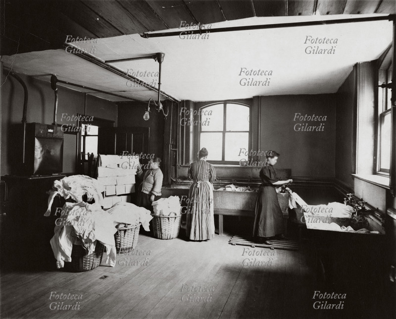 MESTIERI Donne e giovane ragazza al lavoro in una lavanderia. Nel locale sono disposte al centro le ceste di biancheria asciutta e a lato le vasche per il lavaggio ed i risciacqui, la ragazza è addetta al torchio che strizza i panni. Boston, 1905 circa.