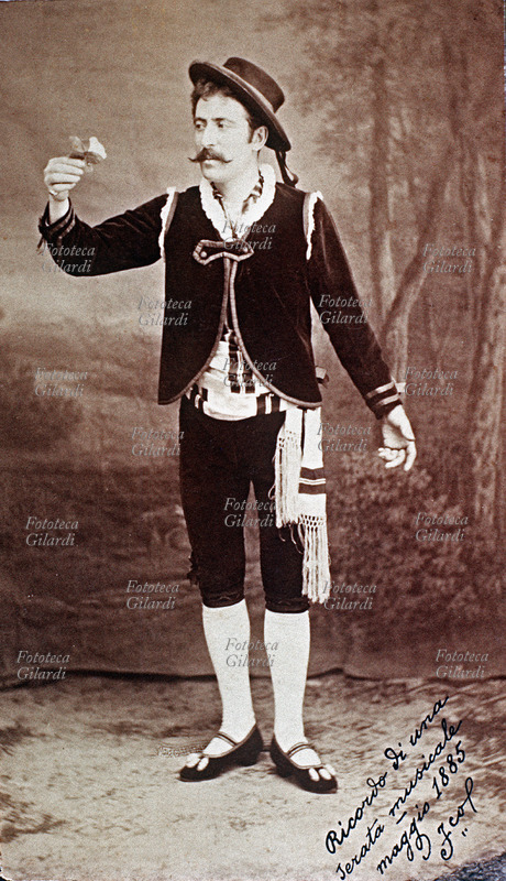 TRAVESTIMENTO Uomo a figura intera in costume spagnolesco, con gesto teatrale osserva intensamente il fiore bianco che tiene nella mano destra, romantico pegno di una dama. Fotografia con dedica "Ricordo di una serata musicale - F.co", Italia, maggio 1885