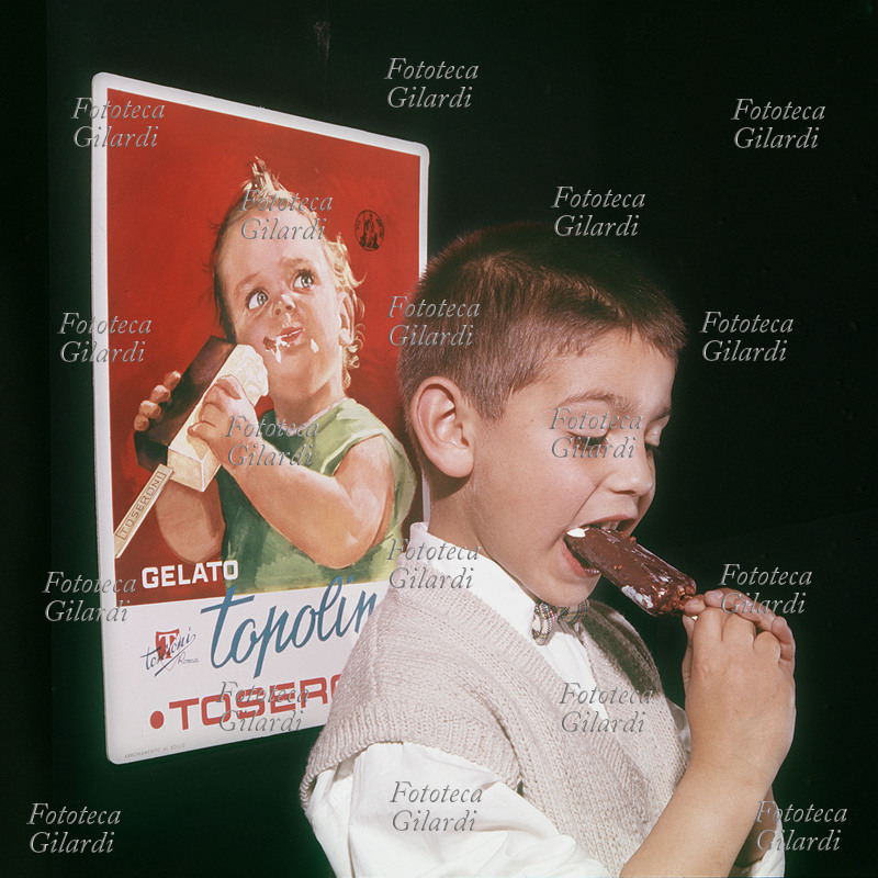 PUBBLICITÀ un bambino mangia il gelato proprio davanti alla locandina pubblicitaria di uno simile al suo. Fotografia di Ando Gilardi (1921-2012) #andogilardi, Italia 1958
