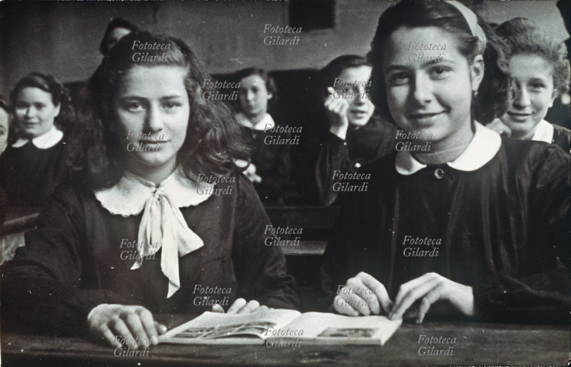 SCUOLA Interno di una classe femminile delle scuole medie inferiori: foto ricordo con la compagna di banco con il sussidiario aperto. Italia, 1952
